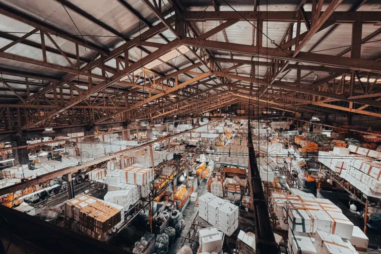 Wide view of a busy warehouse with stacked goods under a steel roof, B2B supply chain