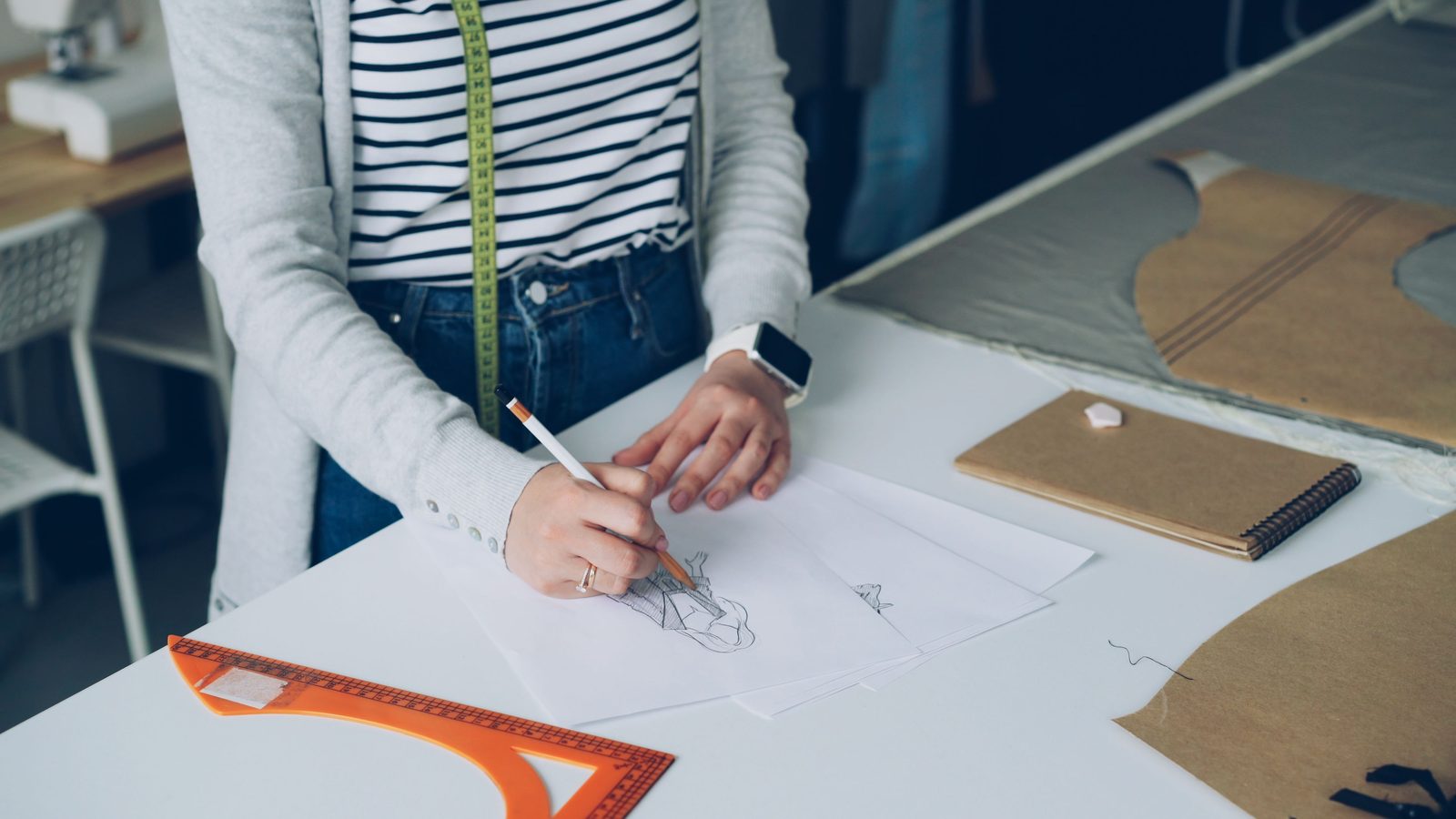 Designer with a tape measure around her neck sketching a garment at a pattern-cutting table, face out of frame