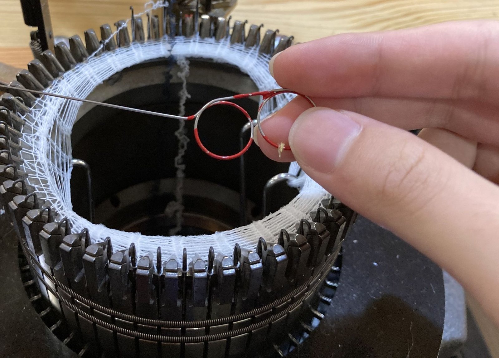 Close-up of needles around a small circular knitting cylinder, with a hand threading white yarn