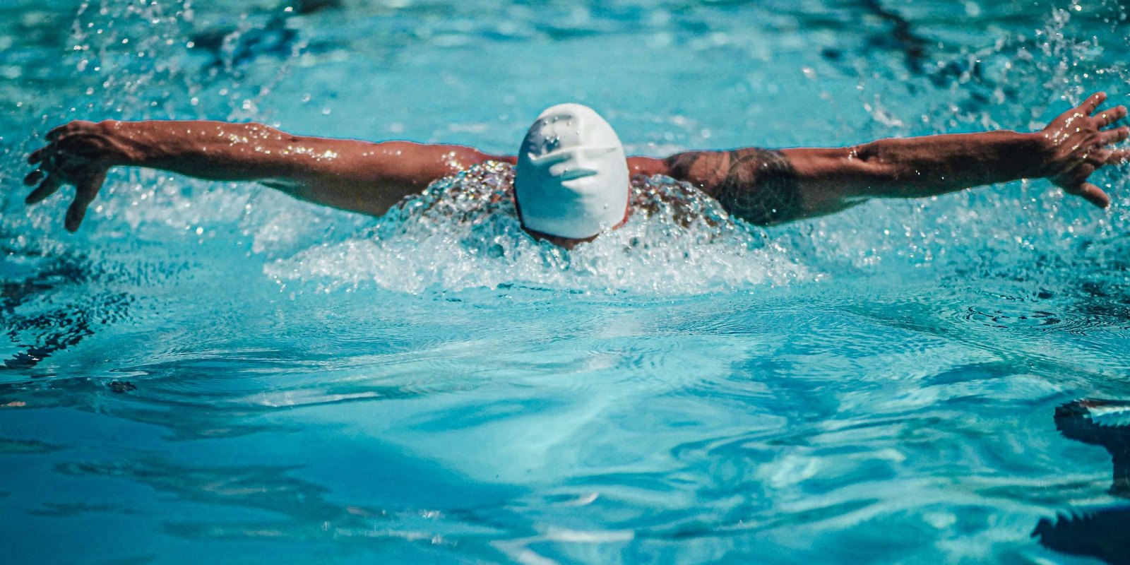 Swimmer in a plain white cap doing butterfly stroke, head down, arms spread above blue pool water